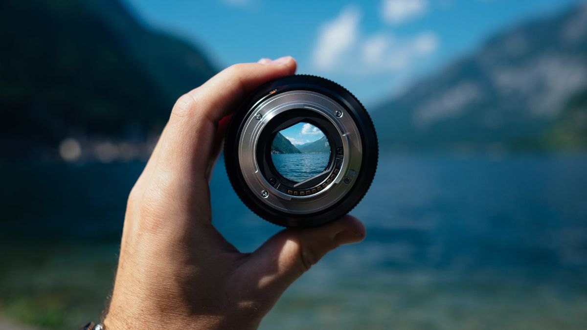 Eine Hand haltet Kamera-Objektiv mit Fokusansicht einer Landschaft mit blauen See